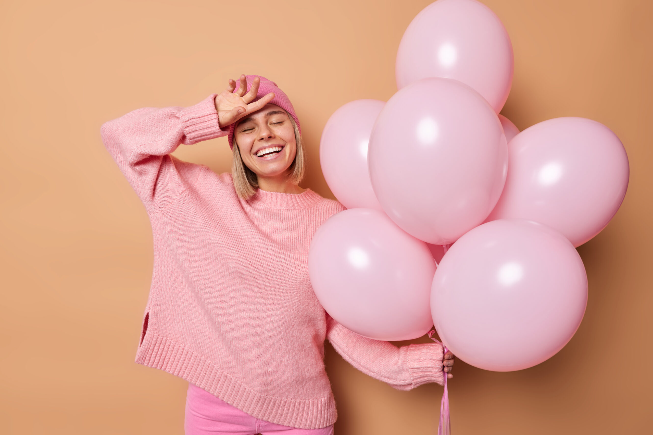 glad carefree young european woman keeps hand forehead smiles broadly has festive mood gets congratulations special event holds bunch inflated balloons isolated brown background 1 scaled